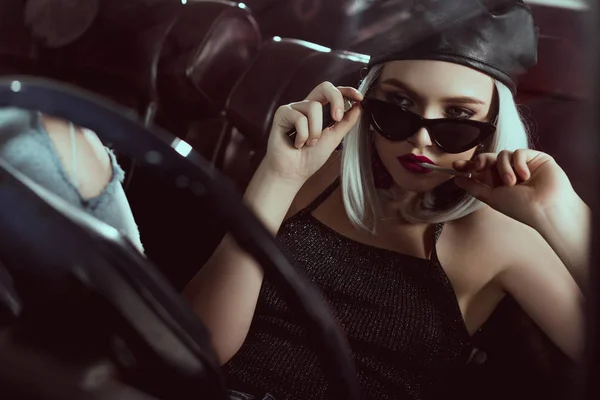 Красива стильна блондинка в береті і сонцезахисних окулярах тримає сигарету, сидячи в машині — стокове фото