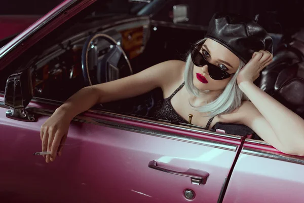Stylische blonde Frau mit Baskenmütze und Sonnenbrille, Zigarette in der Hand und Blick in die Kamera, während sie im Retro-Auto sitzt — Stockfoto