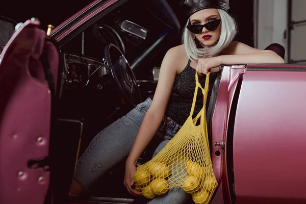 Stylish blonde girl in beret and sunglasses holding string bag with lemons while sitting in car — Stock Photo