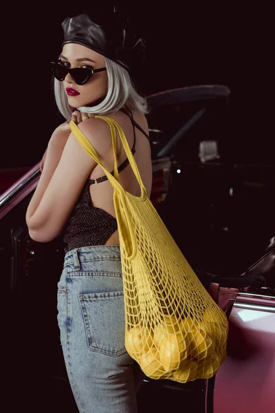 Stylish blonde girl in sunglasses and beret holding string bag with lemons standing near vintage car — Stock Photo