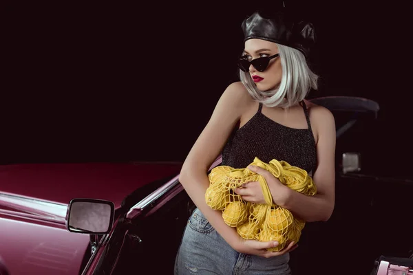 Belle femme blonde en lunettes de soleil et béret tenant un sac à ficelle avec des citrons et regardant loin tout en se tenant près de la voiture — Photo de stock