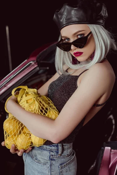Beautiful blonde woman in sunglasses and beret holding string bag with lemons and looking at camera while standing near car — Stock Photo