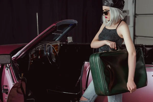 Stylisches blondes Mädchen mit Sonnenbrille und Baskenmütze hält Retro-Koffer in der Nähe von rotbraunem Auto — Stockfoto