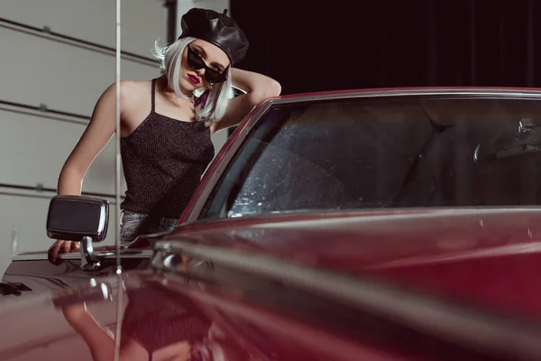 Fashionable blonde woman in black beret and sunglasses leaning at maroon car and looking at camera — Stock Photo