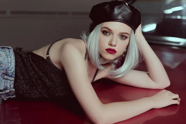 Beautiful blonde woman in black beret lying on maroon car and looking at camera — Stock Photo