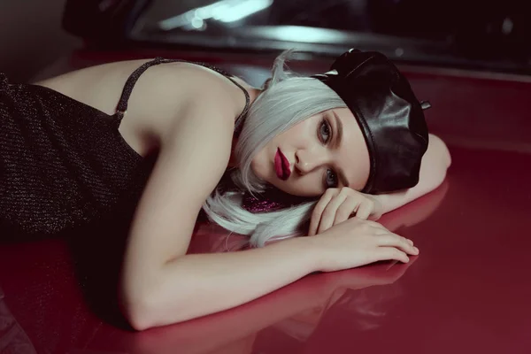 Hermosa mujer rubia en boina negra tumbado en el coche clásico rojo y mirando a la cámara - foto de stock