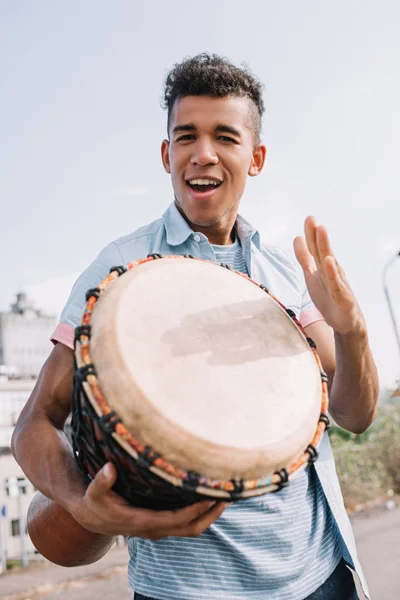 Jeune et heureux musicien de rue afro-américain avec djembe se produisant sur la rue ensoleillée de la ville — Photo de stock