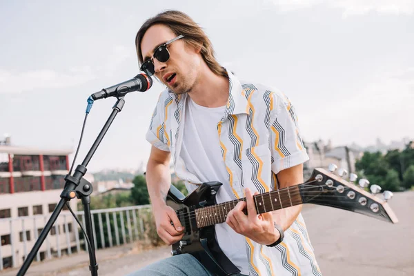 Молодой человек в солнечных очках играет на гитаре и поет в городской среде — стоковое фото