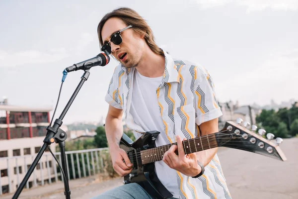 Joven con gafas de sol tocando la guitarra y cantando en ambiente urbano - foto de stock