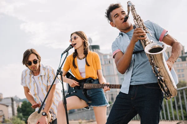 Multiracial young people with guitar, djembe and saxophone performing on street — Stock Photo