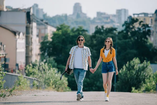 Happy couple of hipster buskers walking and holding hands at city street — Stock Photo