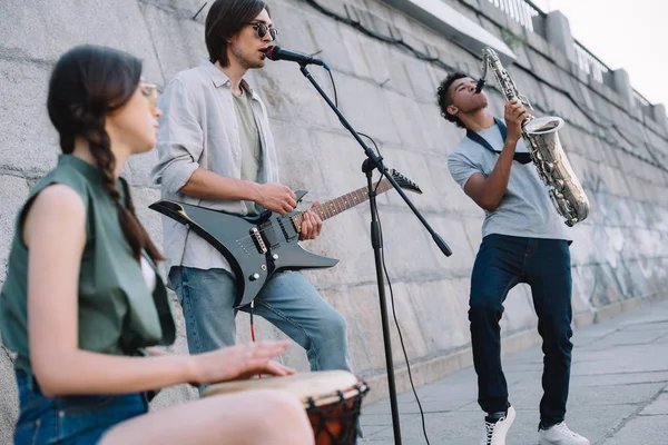 Street musicians band performing with guitar, drum and saxophone on sunny city street — Stock Photo