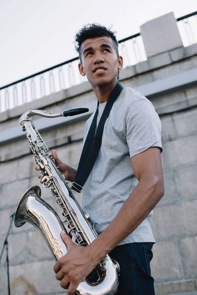 Joven y feliz afroamericano músico callejero sosteniendo saxofón en la ciudad - foto de stock