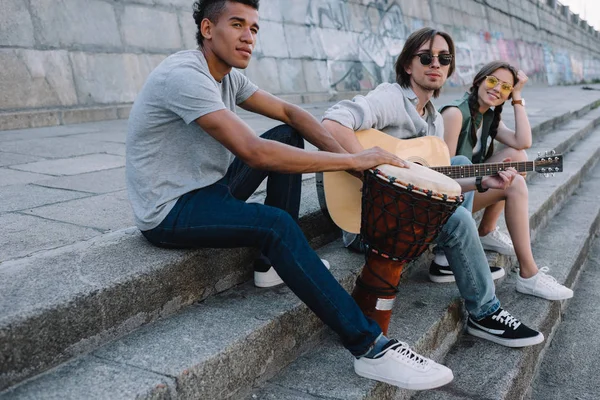 Jóvenes y felices músicos callejeros multirraciales sentados con instrumentos en la ciudad - foto de stock