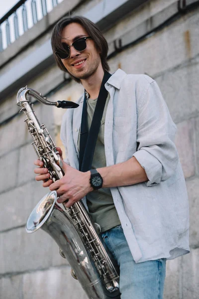Hombre hipster sonriente en gafas de sol sosteniendo saxofón en ambiente urbano - foto de stock