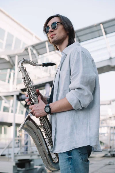 Stylish guy in sunglasses holding saxophone in city — Stock Photo