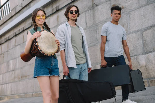 Young girl and multiracial men walking and carrying musical instruments on sunny city street — Stock Photo