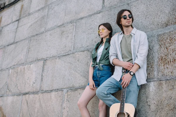 Jeune fille et homme en lunettes de soleil tenant la guitare par mur sur la rue — Photo de stock