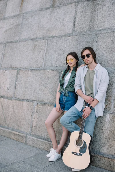 Young and happy couple in sunglasses with guitar by wall in city — Stock Photo