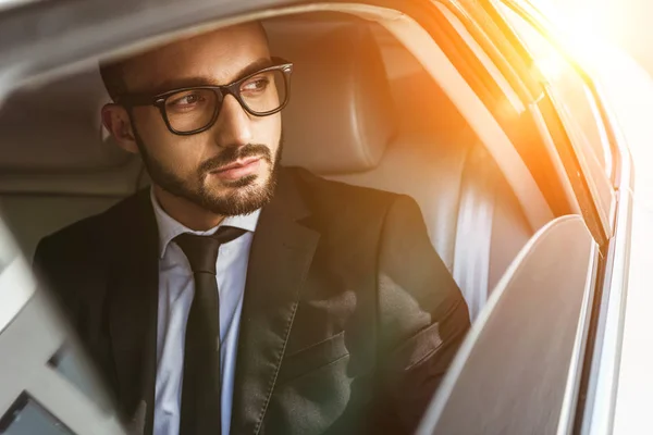 Hombre de negocios guapo sentado en el coche y mirando hacia otro lado durante el atardecer - foto de stock