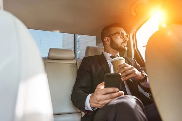 Homem de negócios bonito sentado no carro com copo de café descartável e smartphone — Fotografia de Stock