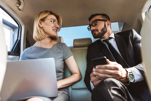Low angle view of handsome businessman and assistant working in car with laptop and smartphone — Stock Photo