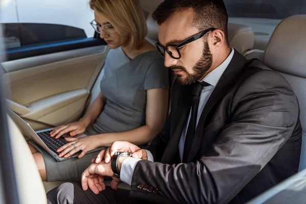 Handsome businessman checking time and assistant working with laptop in car — Stock Photo