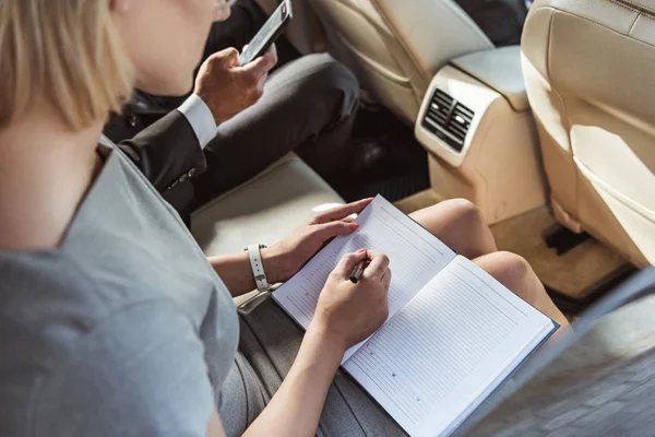 Cropped image businessman and assistant working in car with smartphone and notebook — Stock Photo
