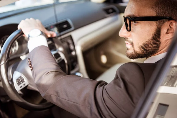 Vue latérale du beau conducteur en costume et lunettes voiture de conduite — Photo de stock