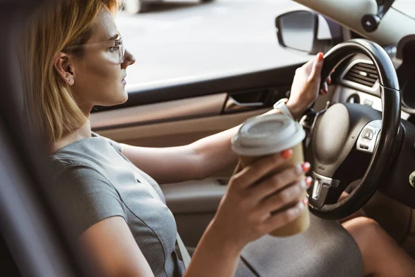 Вид сбоку привлекательной бизнесвумен за рулем автомобиля и держащей кофе в бумажной чашке — стоковое фото