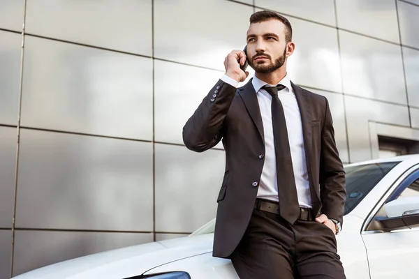 Hombre de negocios guapo hablando por teléfono inteligente y de pie cerca de coche en la calle - foto de stock