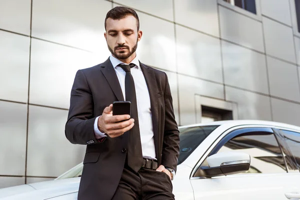 Guapo hombre de negocios sosteniendo teléfono inteligente y de pie cerca del coche — Stock Photo
