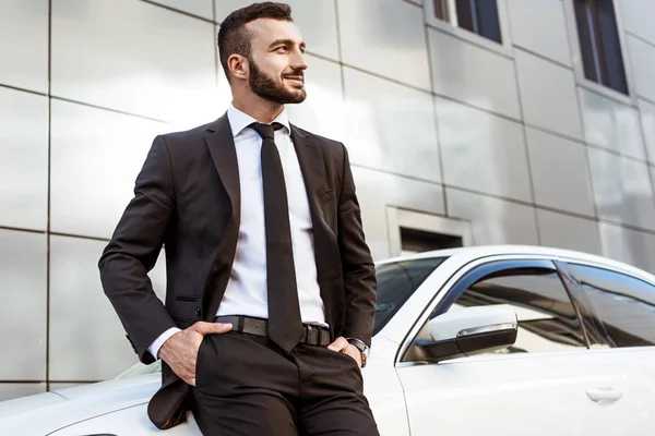 Sorridente bonito empresário inclinando-se no carro e olhando para longe — Fotografia de Stock