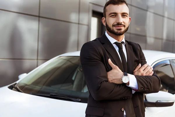 Schöner lächelnder Geschäftsmann, der mit verschränkten Armen neben Auto steht — Stockfoto