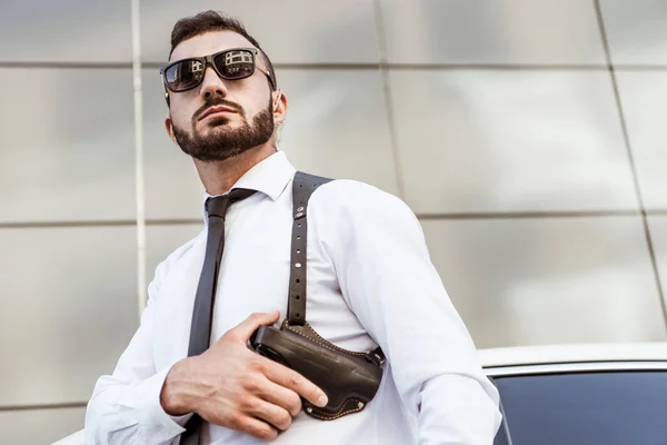 Baixo ângulo de visão de guarda de segurança bonito em óculos de sol tocando arma e olhando para longe — Stock Photo