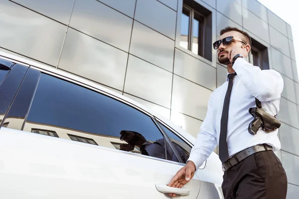 Low angle view of handsome security guard listening message with security earpiece near car — Stock Photo