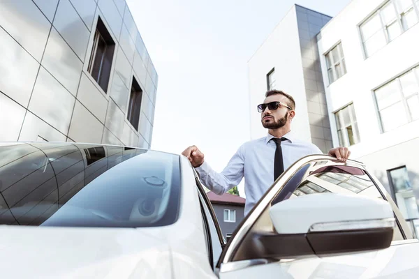 Красивый водитель в солнцезащитных очках стоит рядом с открытой машиной на улице — стоковое фото