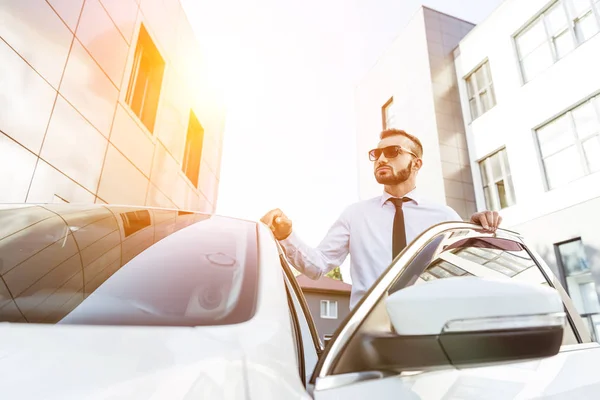 Handsome driver in sunglasses standing near open car on street during sunset — Stock Photo