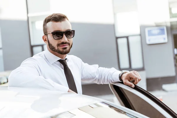 Conductor guapo en gafas de sol de pie cerca del coche y mirando a la cámara - foto de stock