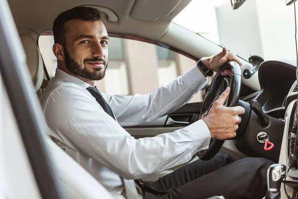 Sourire beau conducteur tenant volant et regardant la caméra dans la voiture — Photo de stock