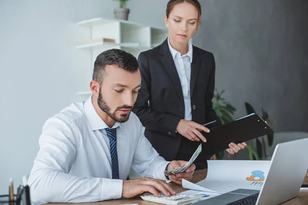 Financier counting money in office, colleague standing with clipboard — Stock Photo