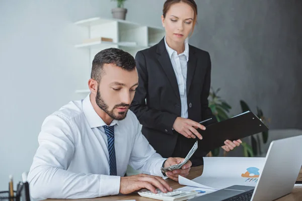 Financiero contando dinero en la oficina, colega de pie con portapapeles - foto de stock