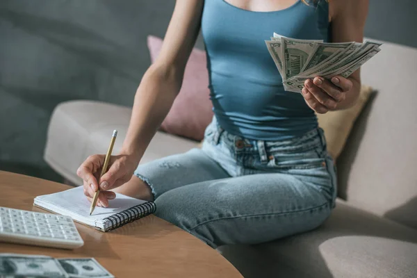 Immagine ritagliata di donna pianificazione di bilancio a casa, in possesso di dollari e la scrittura di qualcosa per notebook — Foto stock