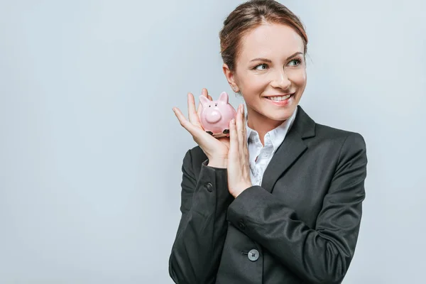 Financiador sorrindo segurando porquinho banco e olhando para longe isolado em cinza — Fotografia de Stock