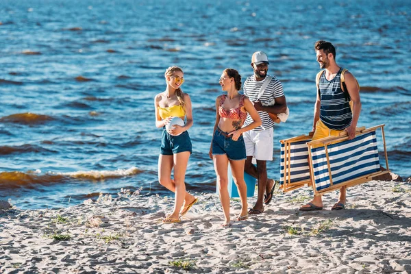 Улыбающиеся молодые многонациональные друзья с пляжными принадлежностями прогуливаясь по песчаному побережью моря — стоковое фото