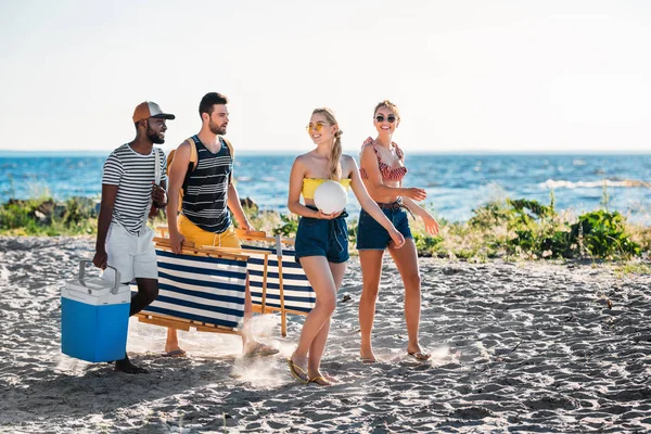 Happy young multiethnic friends with beach chairs, cooler and ball walking on sandy beach — Stock Photo