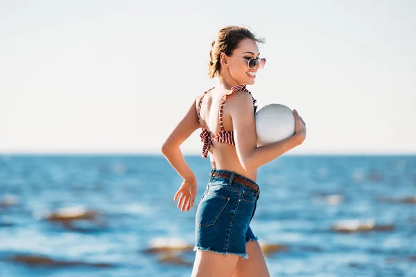 Hermosa mujer joven feliz sosteniendo la pelota y corriendo en la playa - foto de stock