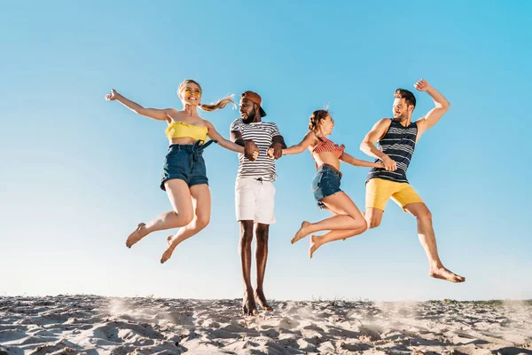 Glückliche junge multiethnische Freunde halten Händchen und springen in Sandstrand — Stockfoto