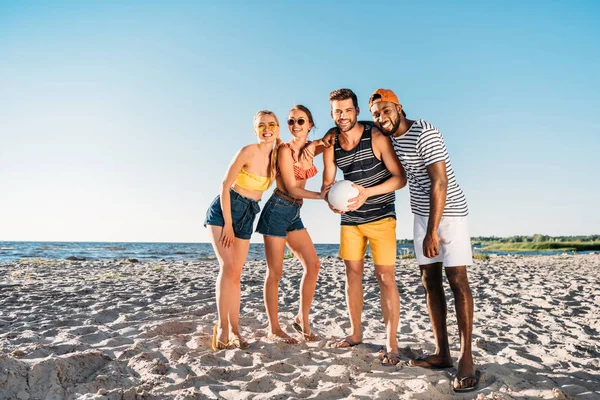 Glückliche junge multiethnische Freunde, die Ball halten und am Strand in die Kamera lächeln — Stockfoto