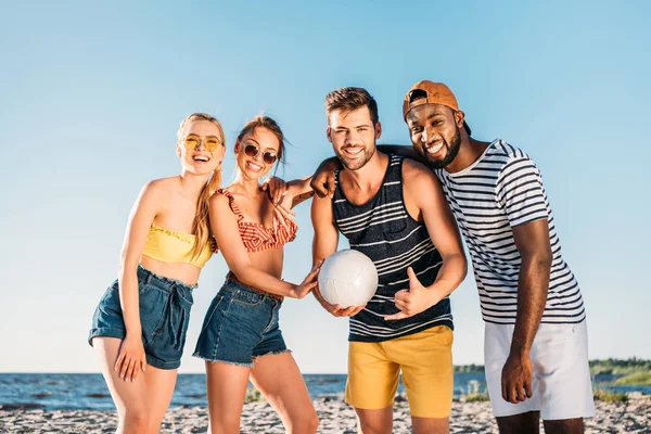 Glückliche junge multiethnische Freunde mit Ball lächeln in die Kamera am Strand — Stockfoto